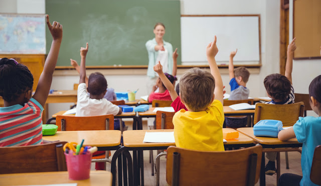 Imagem de crianças, de costas, levantando a mão em uma sala de aula.