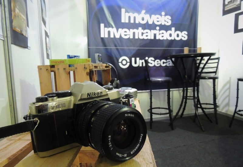Imagem de uma câmera e ao fundo um banner do 'Imóveis Inventariados' da UniSecal.