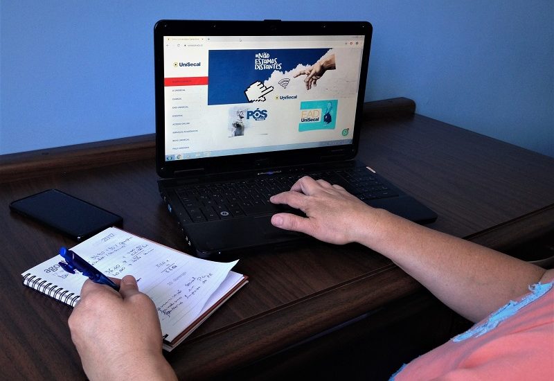 Imagem de uma pessoa utilizando um computador portátil. Ela está no site da UniSecal e fazendo anotações em uma agenda