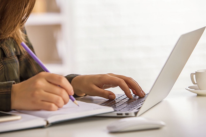 #PraCegoVer: Imagem de uma pessoa fazendo anotações em um caderno. Em frente dela há um computador portátil.