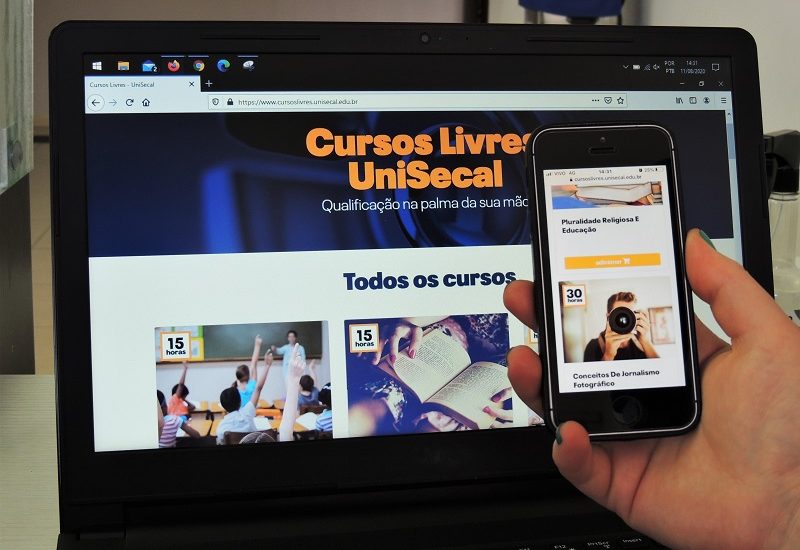 #PraCegoVer: Imagem de uma mão segurando um celular. Na tela do aparelho está o site dos Cursos Livres da UniSecal. Ao fundo há a tela de um computador com o site dos Cursos Livres da UniSecal.