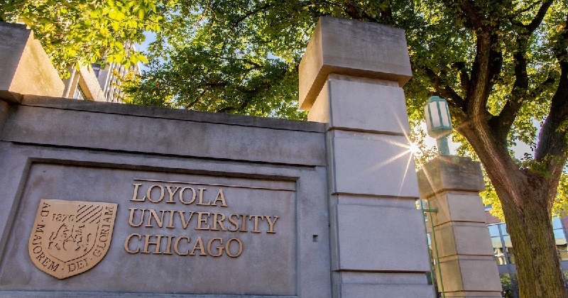 #PraCegoVer: Imagem mostra um muro alto. Ele tem um brasão e o nome da Universidade Loyola de Chicago. Ao seu lado tem uma árvore.