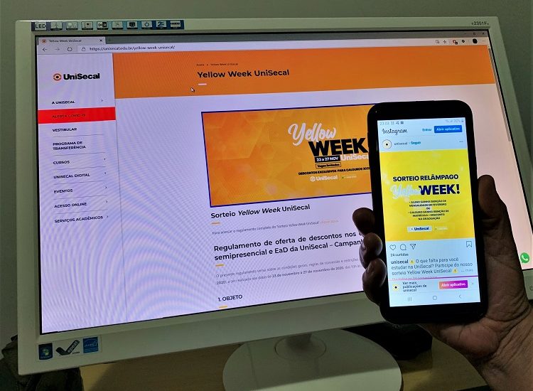 #PraCegoVer: Foto de uma mão, à direita, segurando um celular. Na tela do aparelho está a publicação do 'Sorteio Yellow Week UniSecal'. Atrás há a tela de uma computador e nela está aparecendo a página da 'Yellow Week UniSecal'.