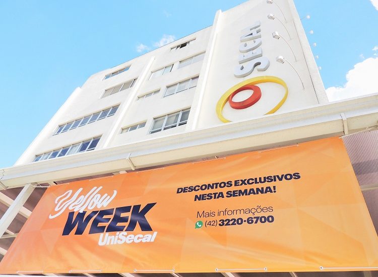 #PraCegoVer: Na foto há o banner da 'Yellow Week UniSecal'. Atrás dele, na parte superior, se encontra o Campus Barão do Cerro Azul da UniSecal.
