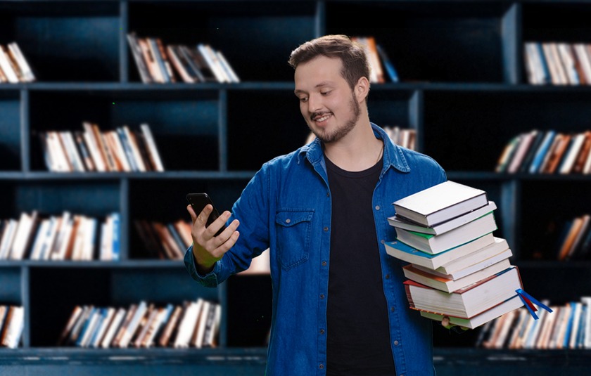 #PraCegoVer: Imagem mostra um estudante dentro de uma biblioteca. Ele segura vários livros em uma mão, e na outra, um celular.