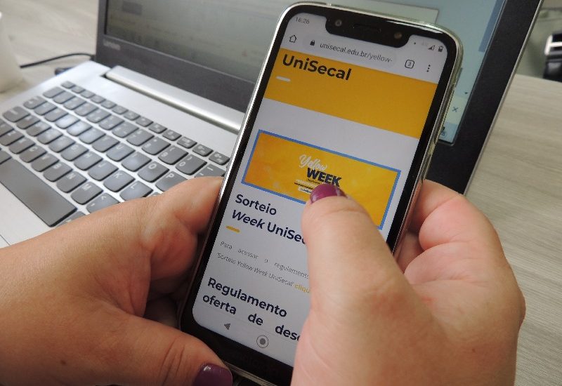 #PraCegoVer: Imagem mostra duas mãos segurando um celular. Na tela está aparecendo o site da UniSecal. No fundo da imagem há um notebook.